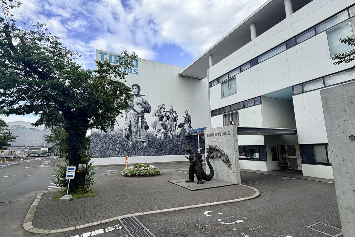 白い建物の門の前。壁に大きな怪獣の絵が描かれ、門の脇にゴジラの像が立っている