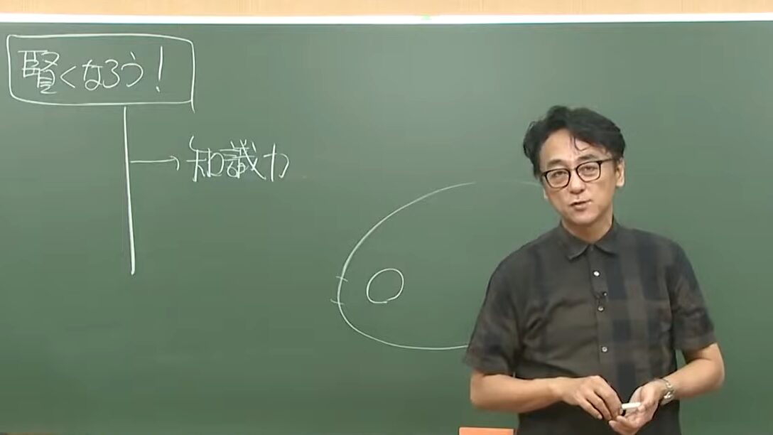 黒板の前で、両手を胸の前に置いて話す青木純二先生。黒板には「賢くなろう!」「知識力」の文字