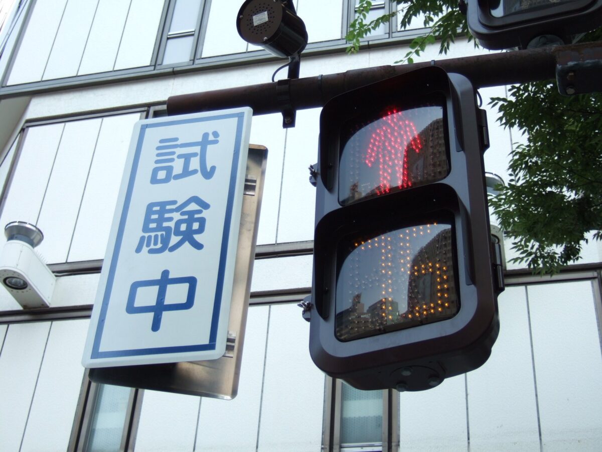 建物の前に下がった歩行者用の信号。赤い人の形が細かい光の粒で光り、横に「試験中」の札がある