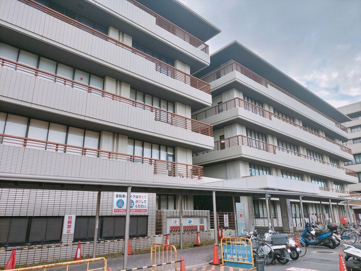 曇り空の下に建つ、ベランダが段々に重なった白い大きな病院の建物。手前に自転車置き場がある