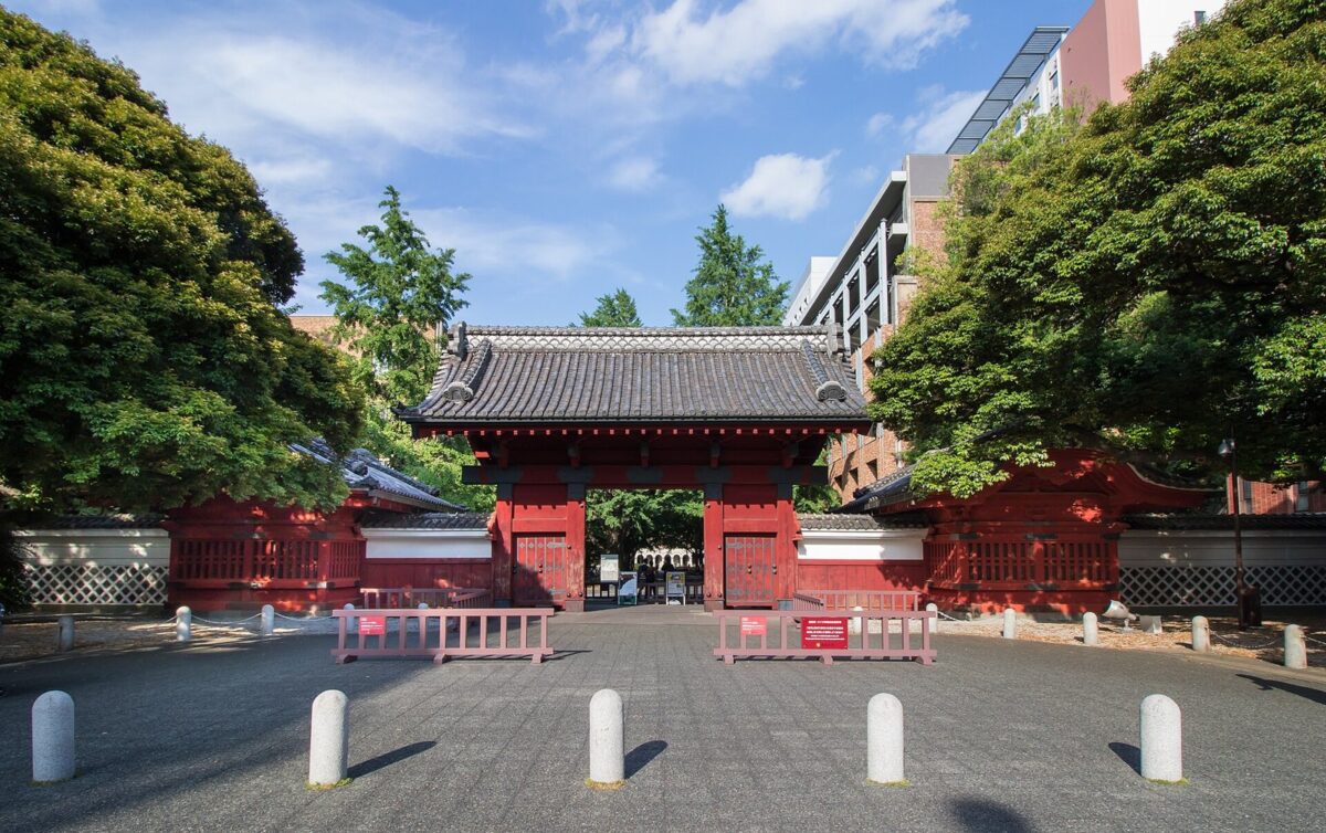 朱色に塗られた瓦屋根の古い門。門の奥に木々と大学の建物が見える