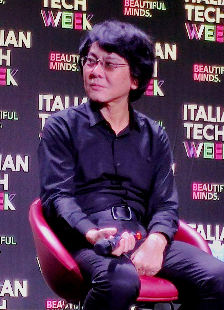 赤い椅子に座り、マイクを手にした黒い服の石黒浩先生