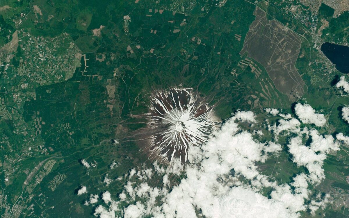 宇宙から見下ろした、頂上に雪の残る富士山。まわりに森と畑が広がり、右下に雲がかかっている