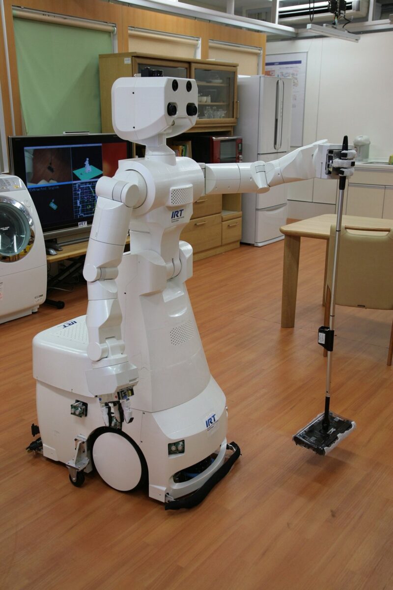 家の台所のような部屋で、車輪のついた白いロボットが片腕を伸ばし、長い柄の道具を持っている
