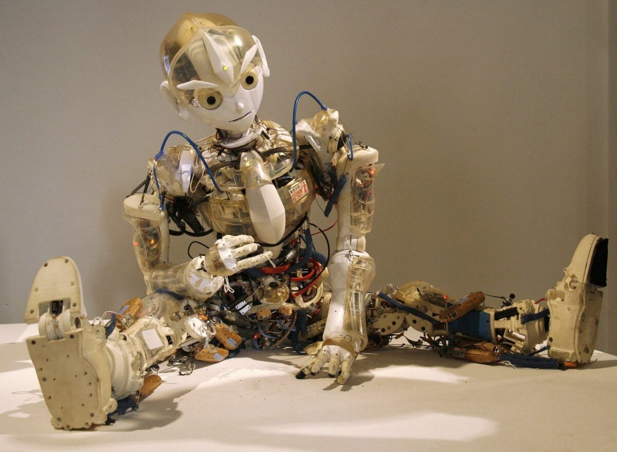 透明な外装の中に配線と部品が見える人型ロボットが、台の上で足を伸ばして座っている
