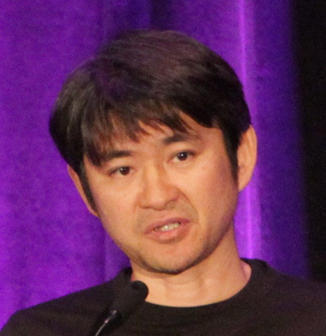 黒い服の男性が、紫の幕の前でマイクに向かって話している