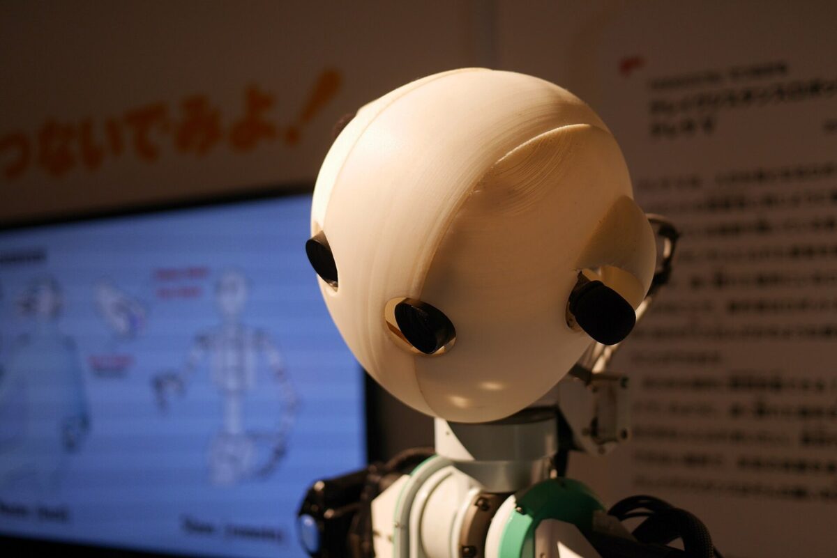 白い丸い頭に黒いカメラの目が2つある人型ロボットの頭と肩。後ろに展示の説明パネルがある