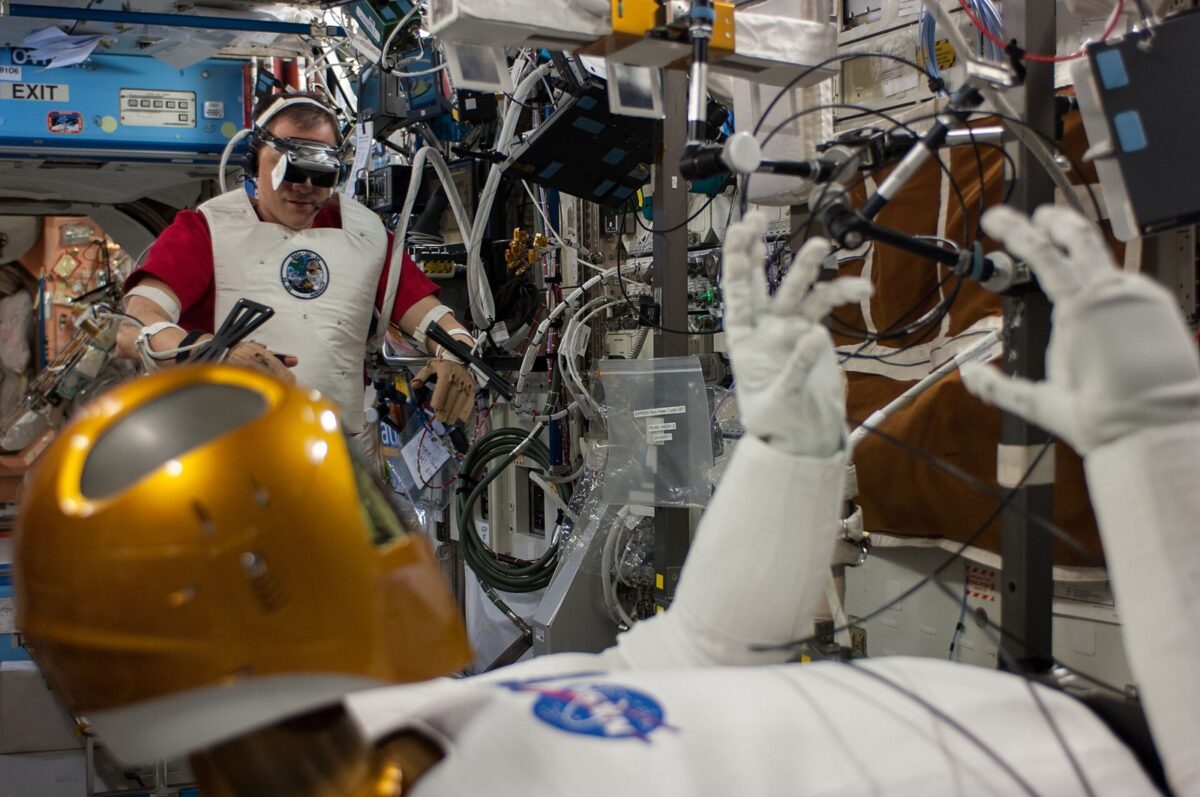 宇宙ステーションの中で、目に装置をつけた宇宙飛行士が両腕を動かし、手前の金色の頭の人型ロボットを操っている