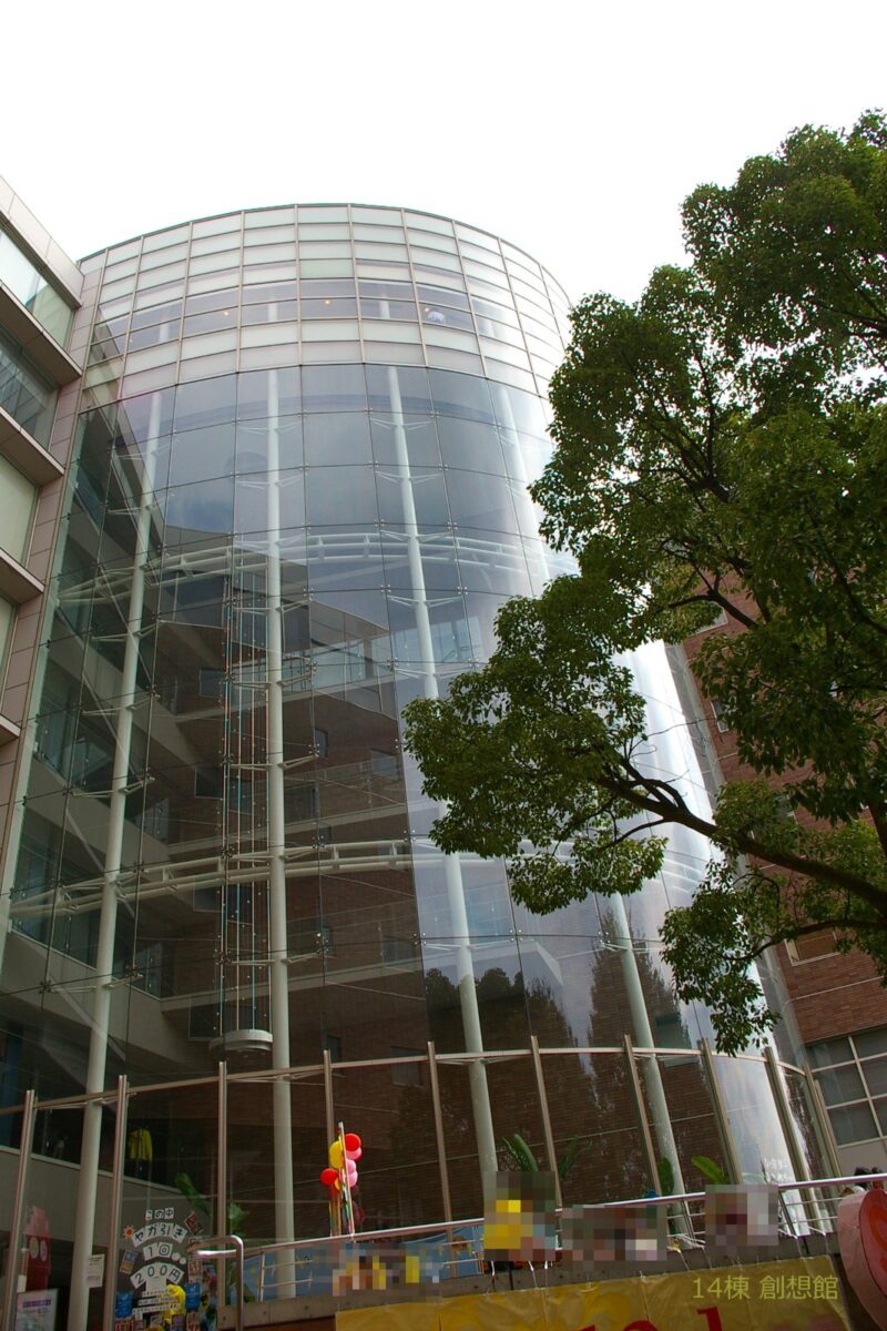 ガラス張りの円い形をした背の高い大学の建物を、木の横から見上げている