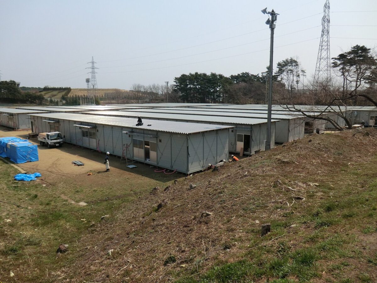 枯れ草の土手の向こうに、灰色の屋根の平屋の仮設住宅が何列も並んでいる。手前に作業用のトラックと青いシートが見える
