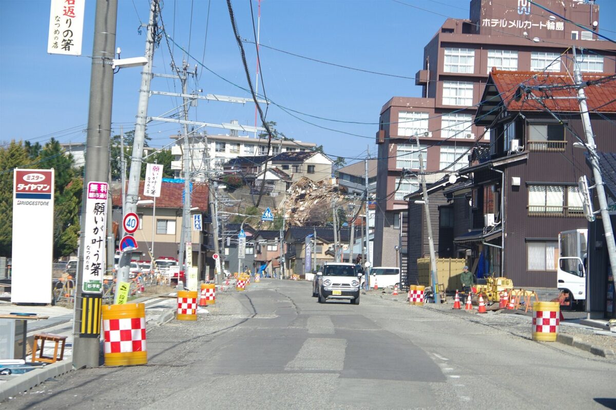 電柱が傾き、道路わきに赤と白の注意の筒が並ぶ町の通り。奥には崩れた建物のがれきが見える