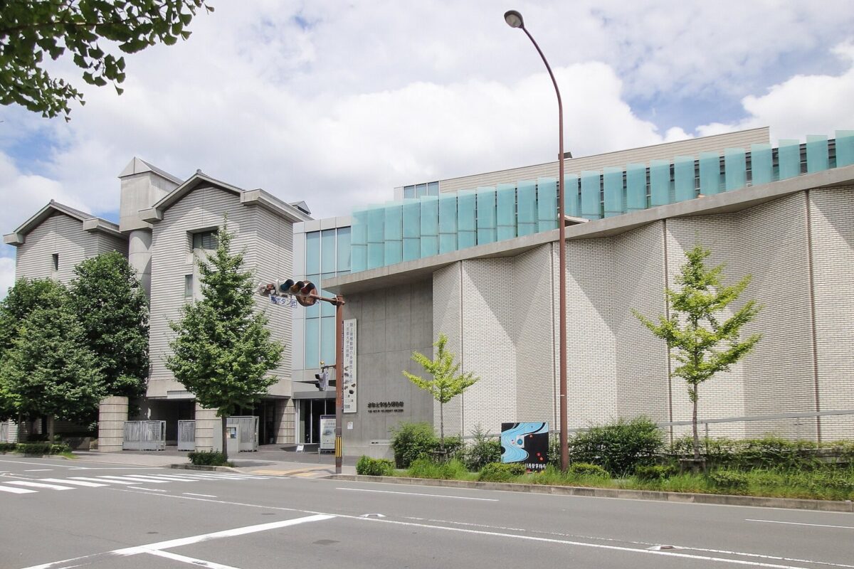 白い壁と青緑のガラスの横長の建物。手前に道路と街路樹がある