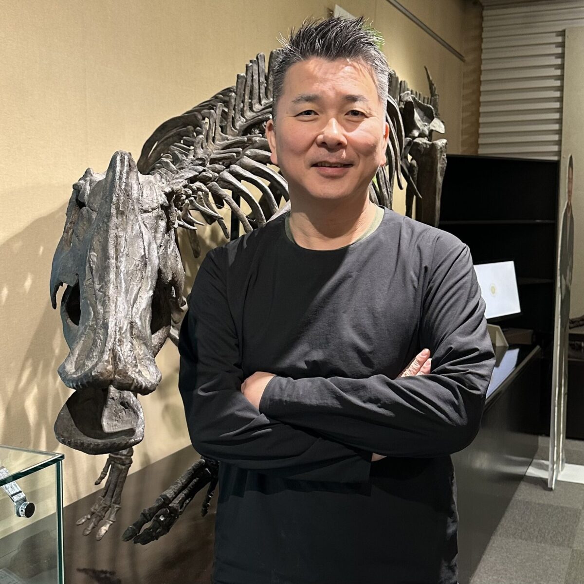 腕を組んでほほえむ黒い服の男性の後ろに、口先の長い恐竜の全身骨格が立っている