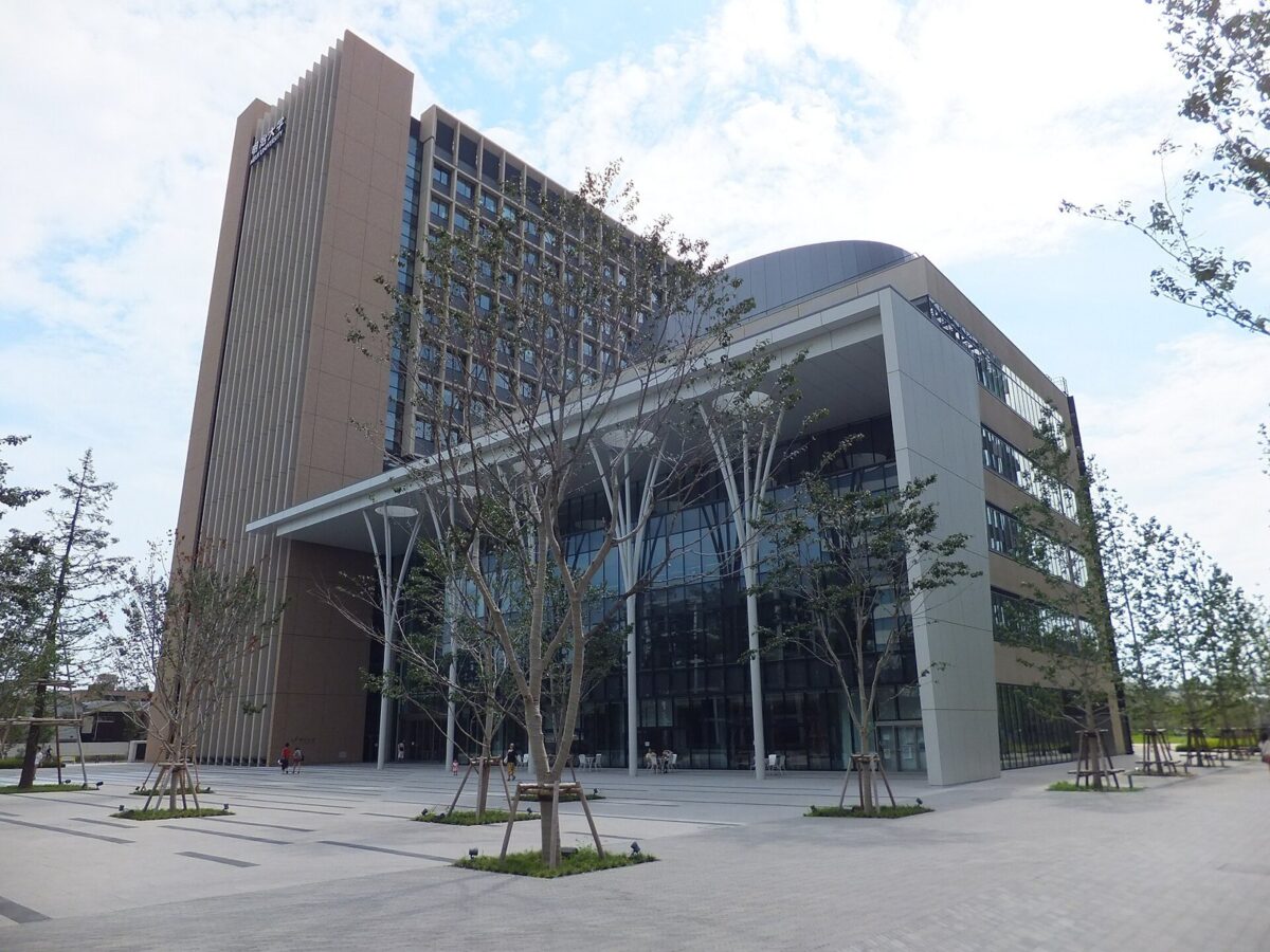 石畳の広場に細い木が並び、奥に高い校舎と、ガラス張りの低い建物が見える