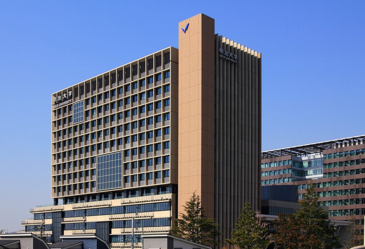 青空の下に建つ、茶色の格子模様の高い校舎。右側に低い建物と木が並んでいる