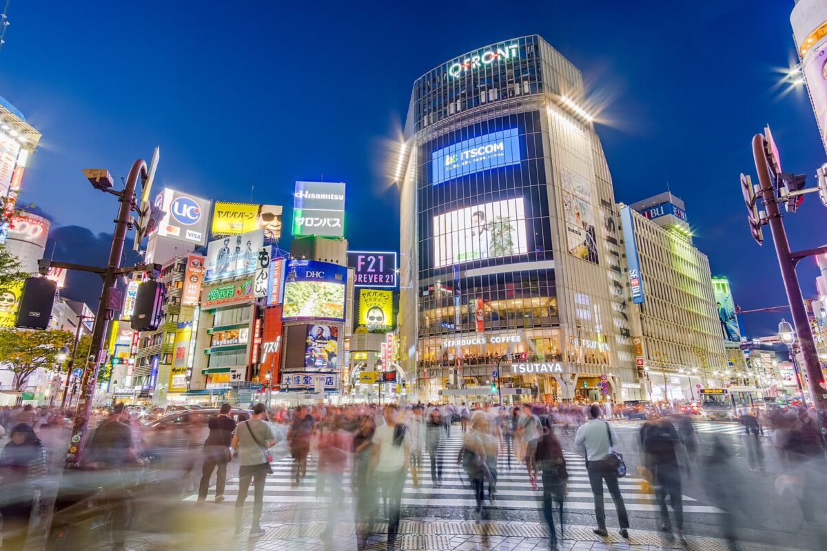 夜の渋谷の交差点。明るい看板のビルの前の横断歩道を、たくさんの人がいろいろな方向へ歩いている
