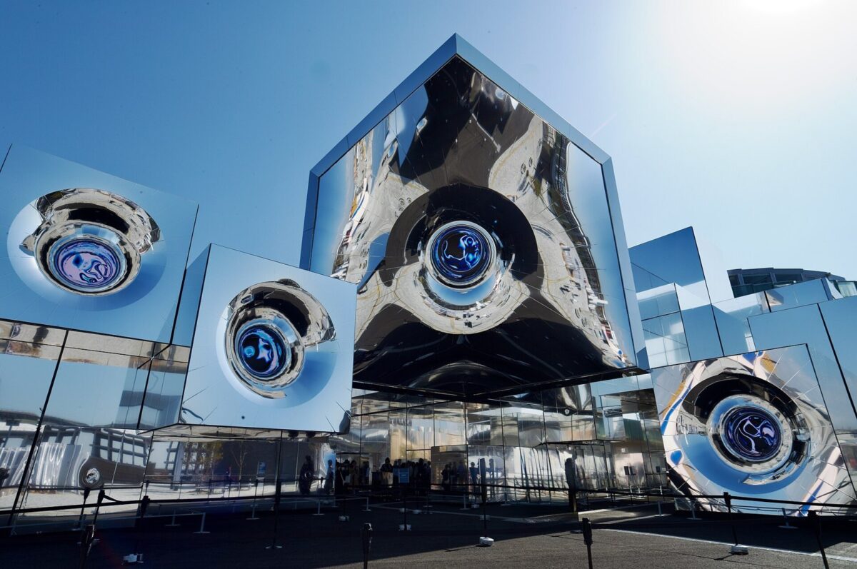 青空の下、鏡でできた大小の立方体が組み合わさった建物。表面が波打ち、周りの景色がゆがんで映っている