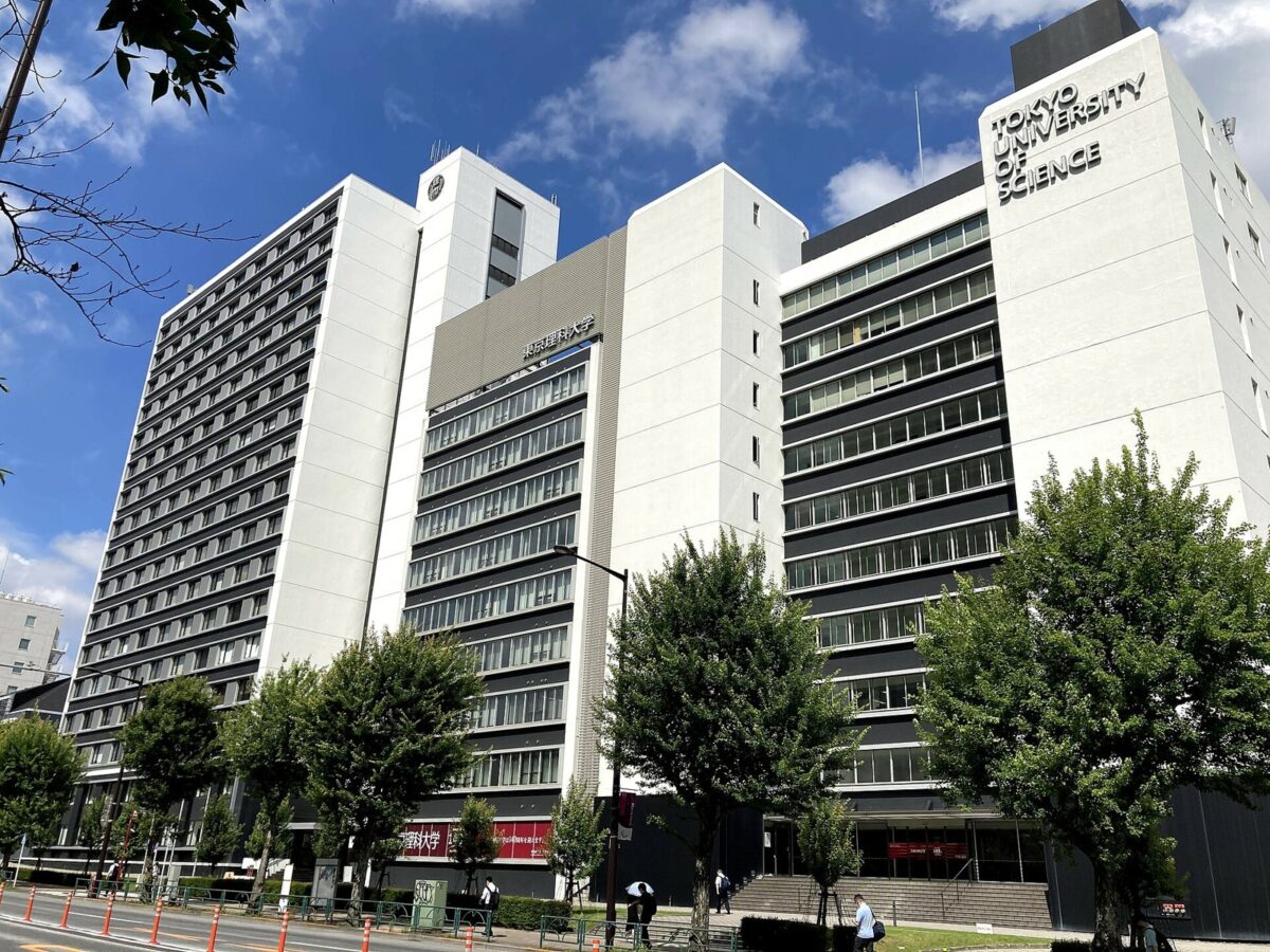 青空の下、大通りに面して建つ白い高層の大学の校舎。上の方に TOKYO UNIVERSITY OF SCIENCE の文字