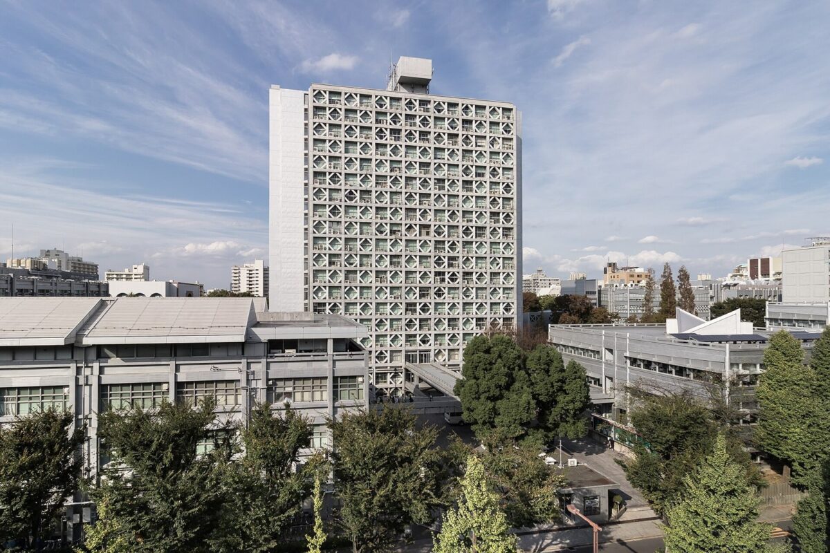 青空の下に建つ、格子もようの窓がびっしり並んだ高い建物。手前に低い校舎と木々がある