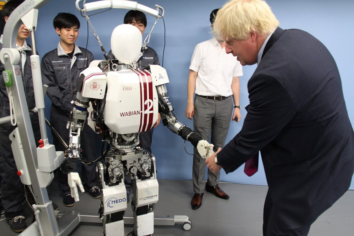 白い人型ロボットの手を、スーツ姿の男性がかがんで握っている。後ろに作業着の学生たちが立っている