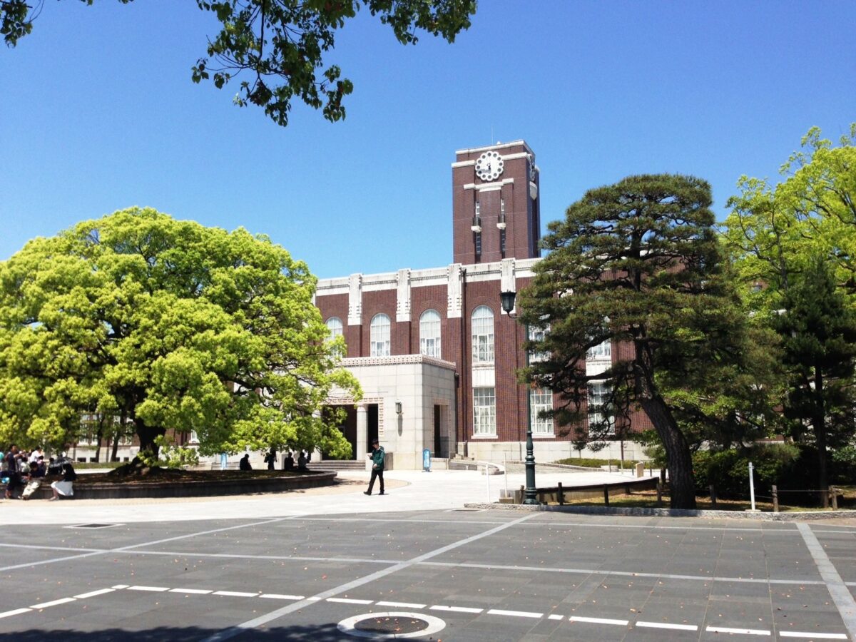 青空の下、赤茶色のれんがの建物の中央に白い時計のついた塔が立っている。左に大きなクスノキ、右に松があり、前の広場を学生が歩いている