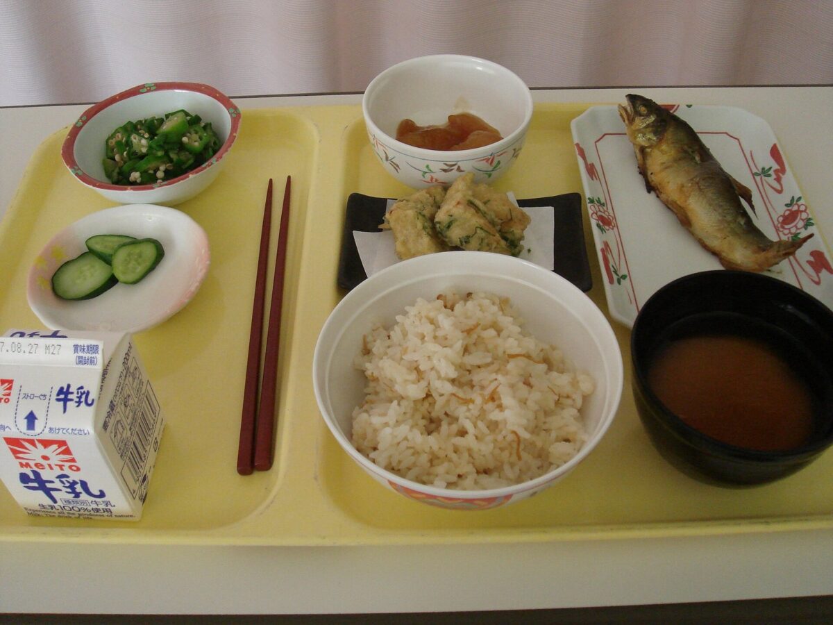 黄色いトレーに、炊き込みごはん、みそ汁、焼いた川魚、天ぷら、きゅうりの酢の物、オクラの小鉢、梅干し、紙パックの牛乳が並んでいる