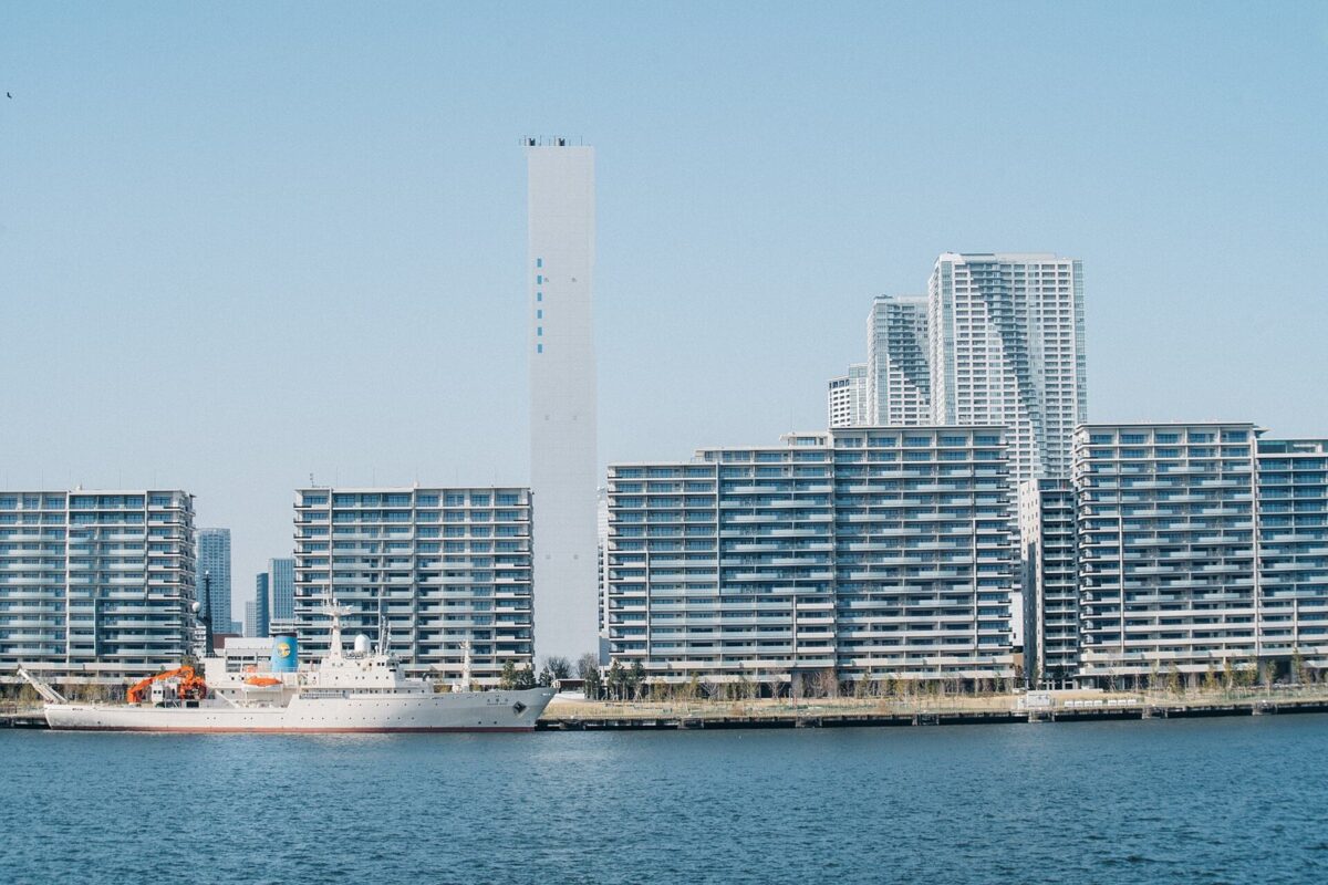晴れた日、海の向こうに、白い大きなマンションの建物がいくつも並んでいる。手前の岸に白い船が止まっている