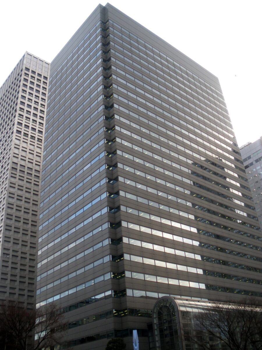 高層ビルが立ち並ぶ街に建つ、灰色の窓が規則正しく並んだ四角い超高層のオフィスビルを、下から見上げた写真