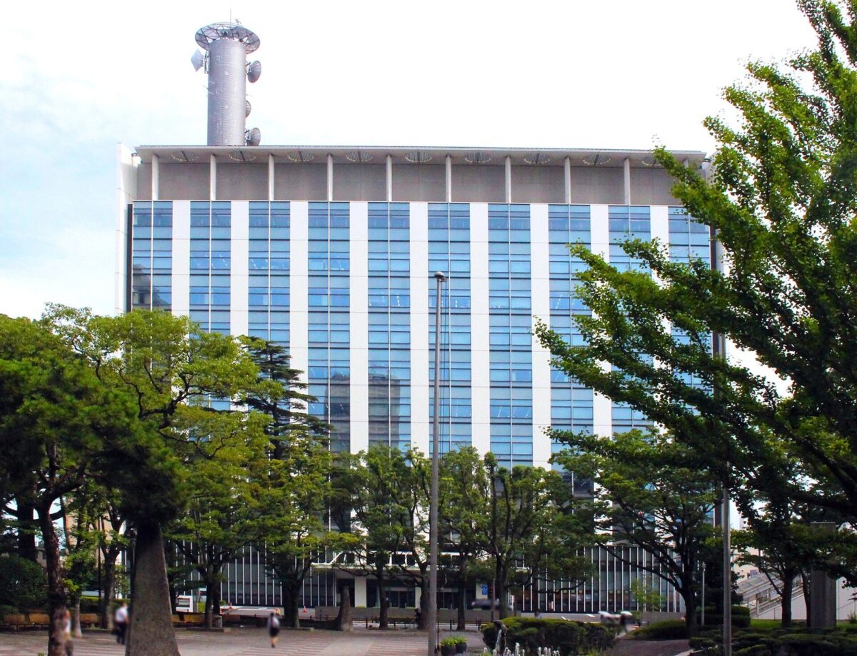 緑の木々の向こうに、青いガラスと白い柱が縦じまに並ぶ高いビルが立ち、屋上に銀色の円柱の塔がある