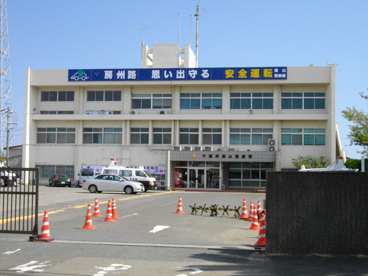 青空の下、白い3階建ての建物の上に青い横断幕があり、手前の駐車場にパトカーとオレンジのコーンが並んでいる