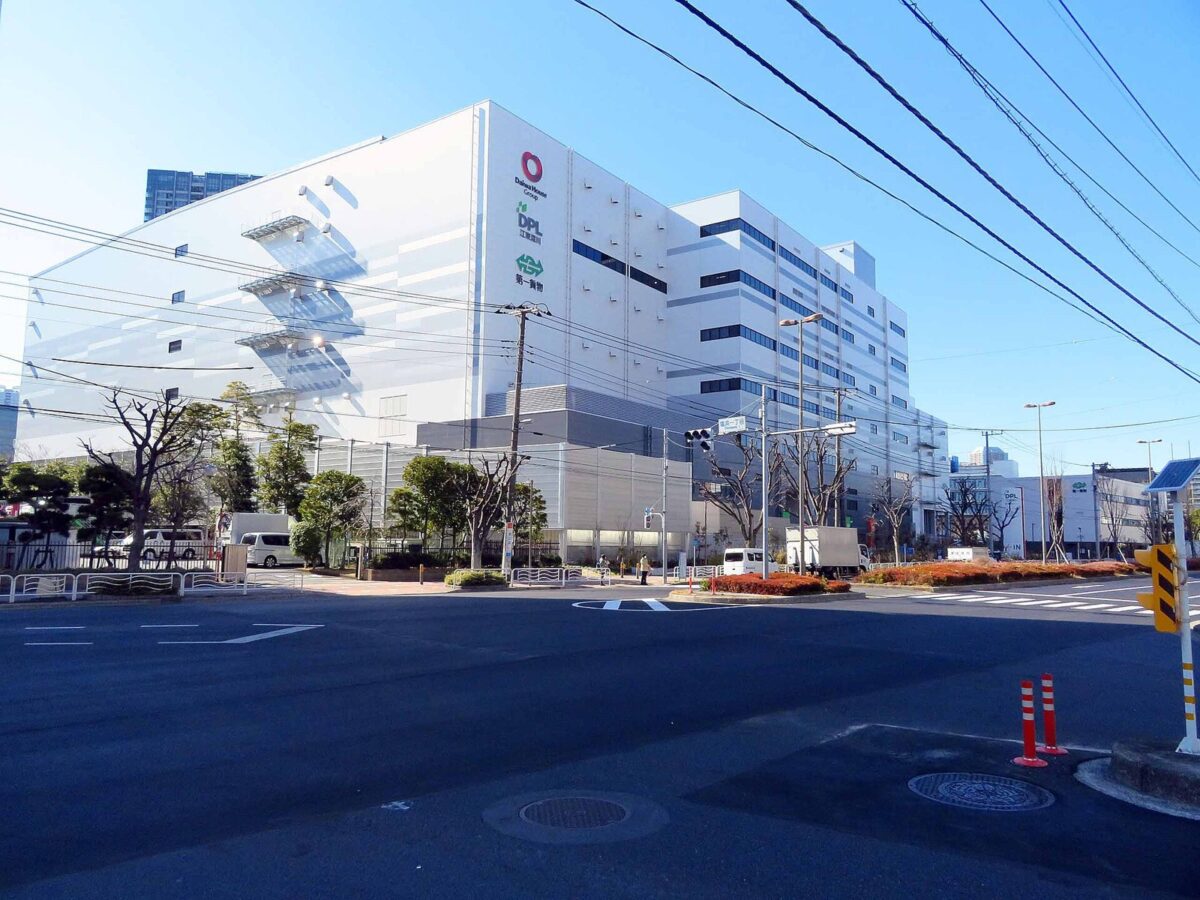 晴れた日の交差点の向こうに建つ、白くて大きな何階建てもの物流倉庫。手前に広い道路と電線がある