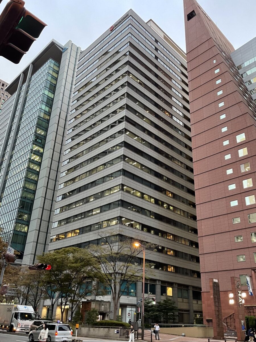 夕方の交差点から見上げた、横じまの窓が並ぶ高層ビル。右に赤茶色の高いビル、足もとに木と車がある