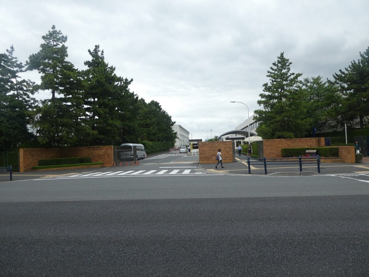 松の木に囲まれた門と、その奥に続く白い建物。手前は広い道路
