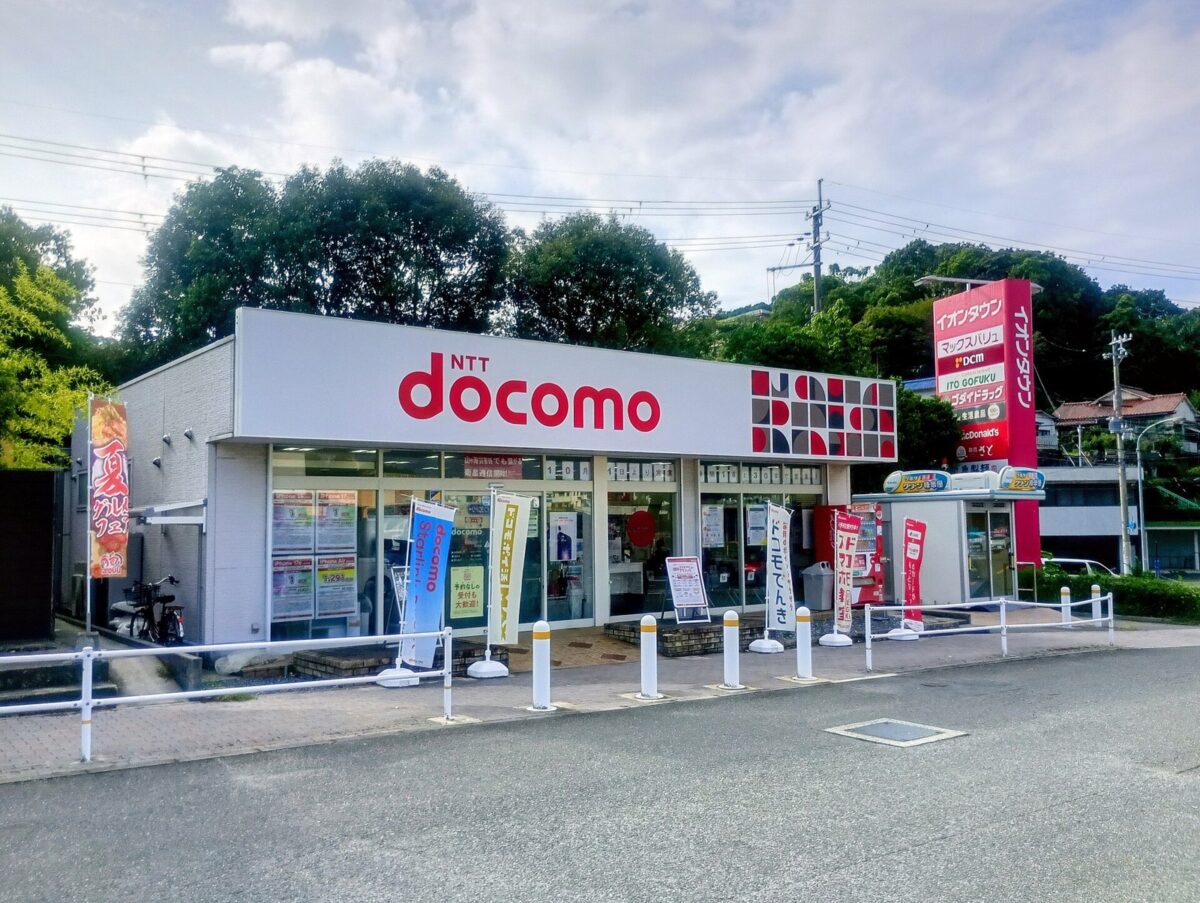 駐車場に面した白い平屋のドコモショップ。赤い「NTT docomo」の看板と、のぼり旗が並ぶガラス張りの入口