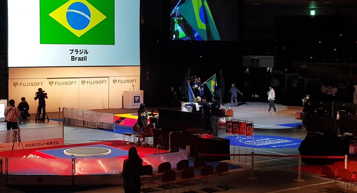 暗い大きな会場で、ブラジルの国旗を持った選手たちが入場している。手前には円い土俵、奥の壁には富士ソフトの名前