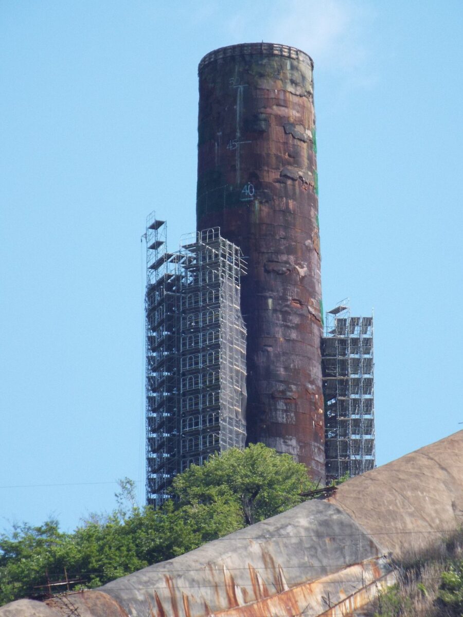 青空の下、緑の山の上に立つ、赤茶色の太い円筒の煙突。まわりに工事の足場が組まれている