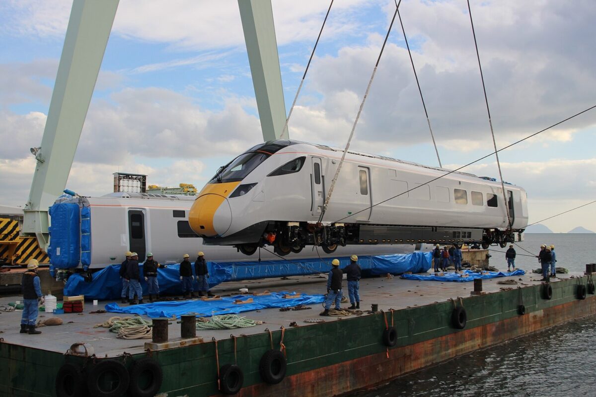 海辺の大きなクレーンにつり上げられ、船の上に下ろされる、先頭が黄色の白い電車。ヘルメットの作業員たちが見守っている