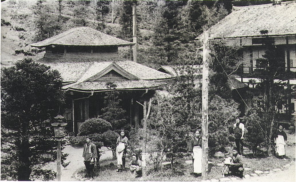 林の中に建つ木造の温泉旅館の建物と、庭に並ぶ着物に前掛けの人たちの白黒写真