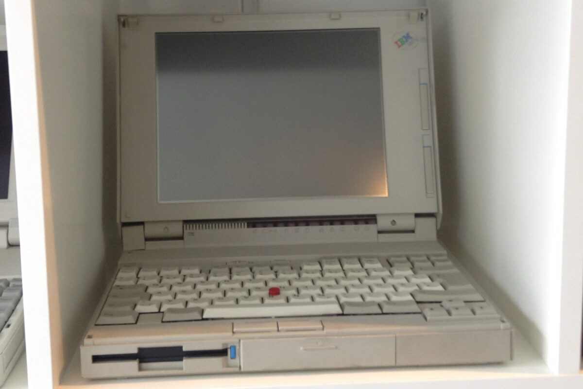 展示の棚に開いて置かれた、灰色の古いノートパソコン。キーボードの真ん中に赤い小さな点がある