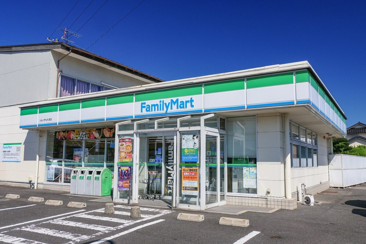 郊外の道沿いにある平屋のファミリーマートの店。白い壁に緑と青の帯の看板、広い駐車場が見える