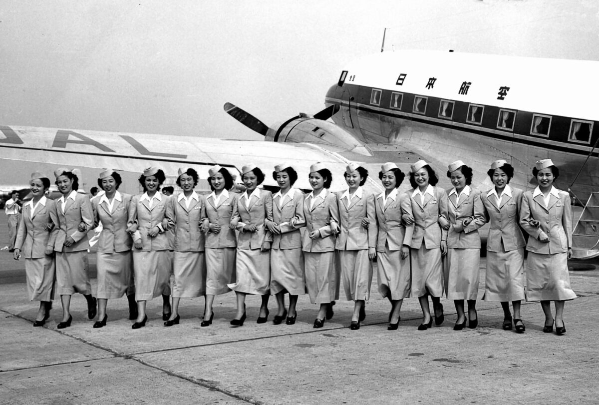 「日本航空」と書かれた古いプロペラ機の前を、制服と帽子の若い女性の客室乗務員15人が腕を組んで一列に歩いている白黒写真