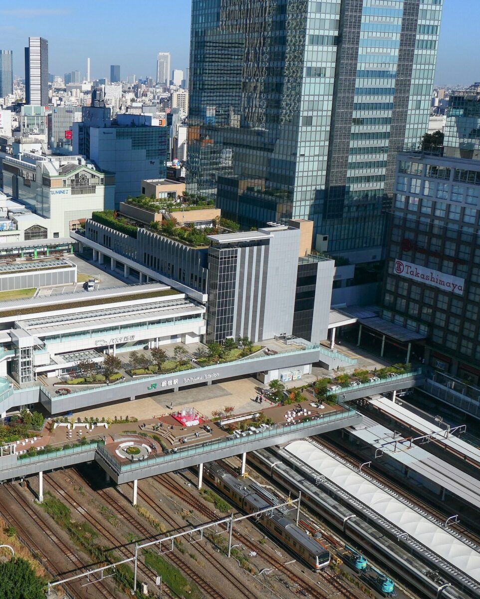 高いビルの上から見下ろした大きな駅。何本もの線路の上に、屋上に緑のある駅ビルと広い広場がかぶさり、奥に高層ビルが並ぶ