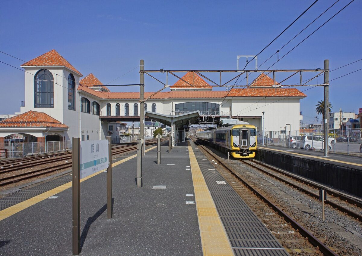 青空の下、白い壁にオレンジ色の瓦屋根の駅舎と、黄色い線のあるホーム。線路には先頭が黄色の列車が止まっている