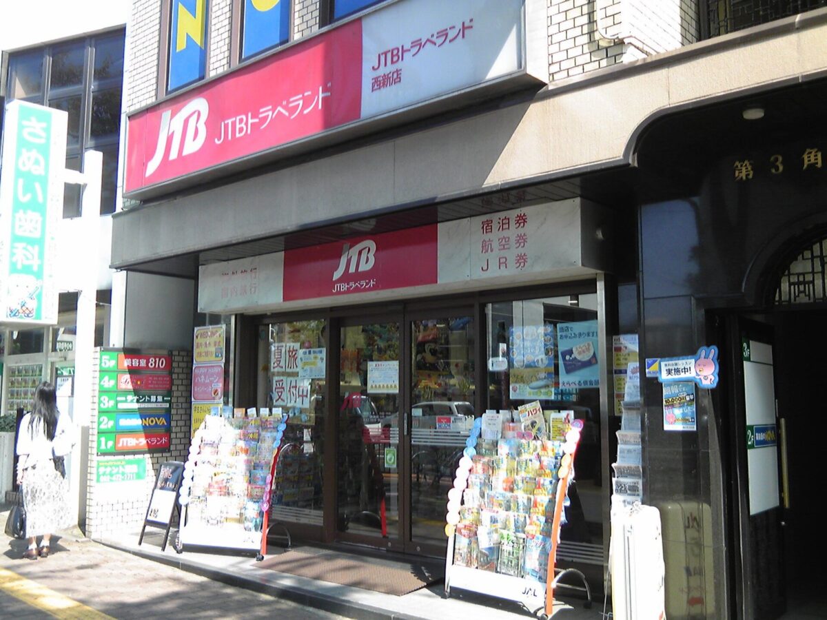 ビルの1階にある旅行会社の店。赤い看板に「JTBトラベランド」、入口の上に「宿泊券 航空券 JR券」、店の前にはパンフレットの棚が並ぶ