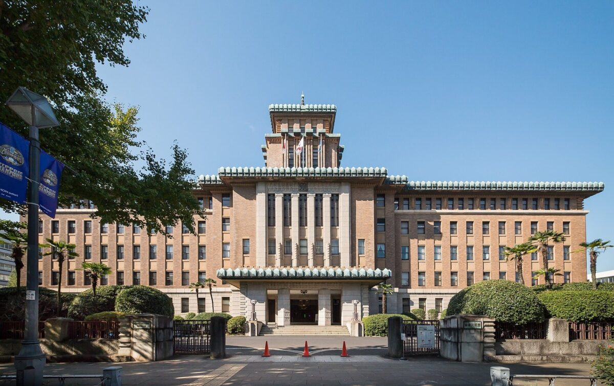 青空の下、真ん中に緑の屋根の塔が立つ、茶色いれんがの横に長い庁舎。手前に門と植え込み