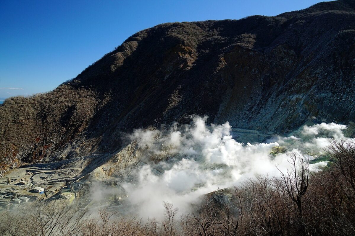 青空の下、木の少ない茶色い山の斜面のふもとから、白い湯気がもくもくと立ちのぼっている