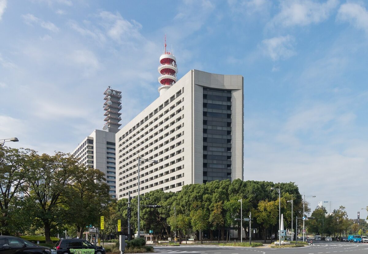 青空の下、上が扇の形に広がった薄いベージュの高層ビル。屋上に赤と白のしま模様の塔があり、手前に緑の木々と広い道路