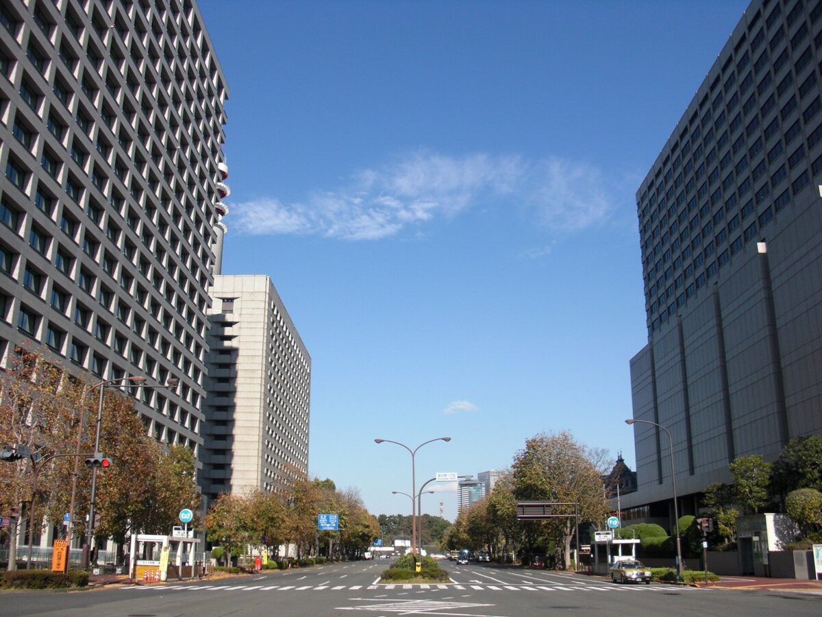 青空の下、広い道路の両側に、窓がびっしり並ぶ大きな役所のビルが建っている