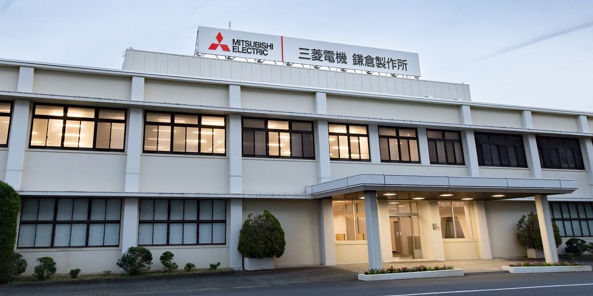 夕方の空の下、屋上に「三菱電機 鎌倉製作所」の看板が立つ、白い2階建ての横に長い建物。窓に明かりがともり、玄関にひさしがある