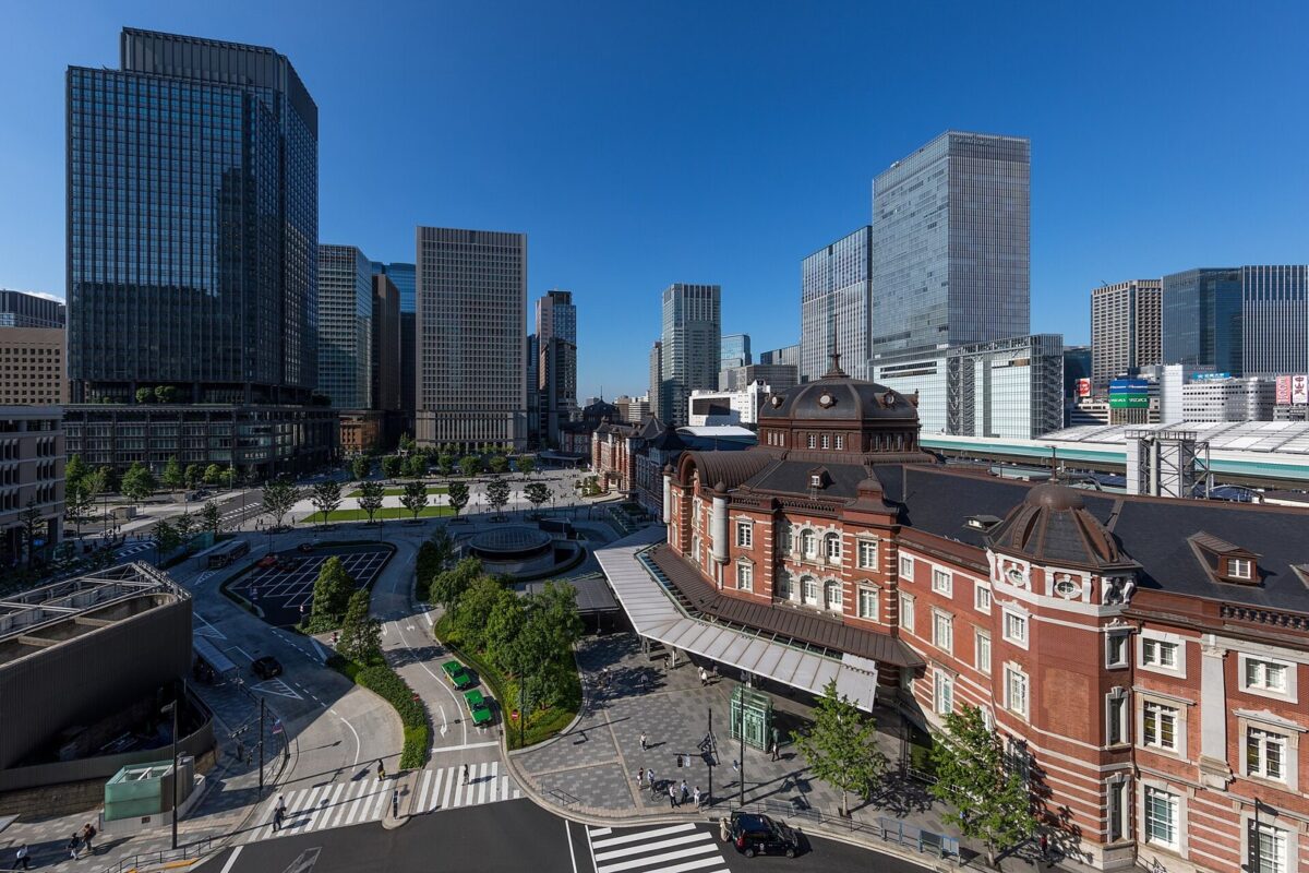 青空の下、赤れんがの東京駅の駅舎と駅前の広場。その奥と左右に、ガラス張りの高層ビルが立ち並んでいる